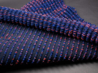 Nahaufnahme eines strukturierten Schlingenstoffs in Dunkelblau mit roten und hellblauen Akzenten, dargestellt auf einer dunklen Oberfläche.