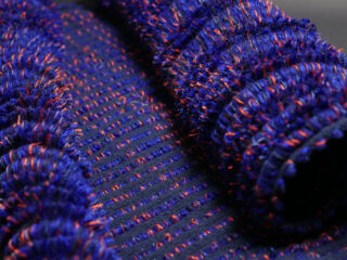 Nahaufnahme eines strukturierten blau-schwarz gewebten Textils mit roten Akzenten und erhabenen, gerüschten und geschlungenen Mustern.