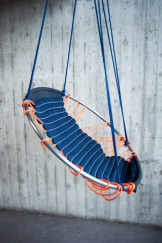 Ein runder Hängesessel mit Metallrahmen, blauer Sitzfläche und orangefarbenen Seilakzenten hängt an blauen Seilen vor einer Betonwand.