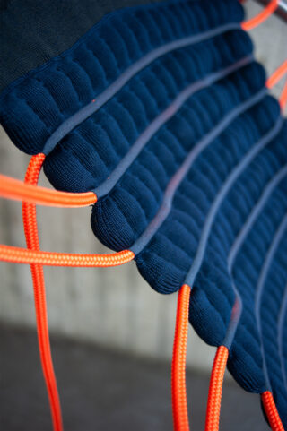 Nahaufnahme einer Stuhlsitzfläche mit dunkelblauen gepolsterten Stoffabschnitten und leuchtend orangefarbenen Schnüren, die die Struktur bilden.