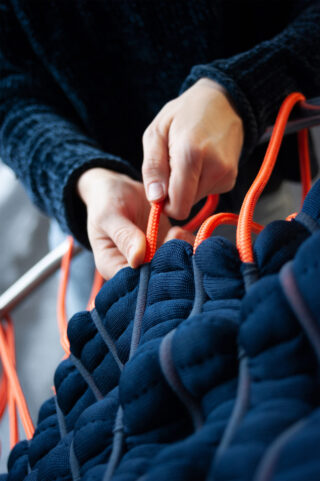Eine Person webt mit ihren Händen ein leuchtend orangefarbenes Seil durch Falten eines dunkelblauen Stoffes.