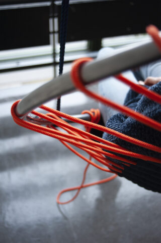 Nahaufnahme von leuchtend orangefarbenen Seilen, die um eine Metallstange geschlungen sind und einen Hängesessel tragen, in dem eine Person im Innenbereich sitzt.