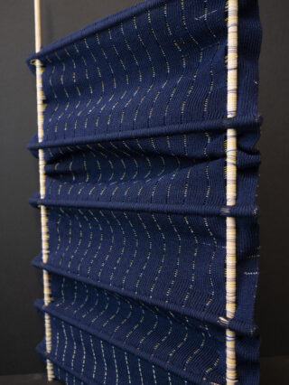 Eine vertikale Textilstruktur aus marineblauem Gewebe, gespannt zwischen parallelen Holzstäben, mit gleichmäßig verteilten horizontalen Falten und kleinen Punktmustern.