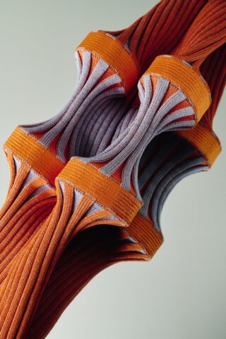 Eine Nahaufnahme von aufwendig gedrehten und gespannten orangefarbenen und grauen gerippten Stoffbändern vor einem neutralen Hintergrund.