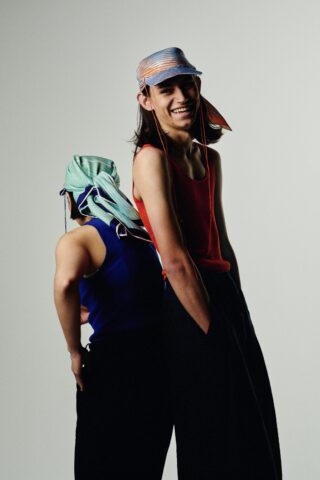 Zwei Personen stehen Rücken an Rücken. Eine blickt nach vorne, lächelt und trägt ein rotes Tanktop und ein gestreiftes Kopftuch. Die andere blickt abgewandt und trägt ein blaues Tanktop und ein grün gestreiftes Kopftuch.