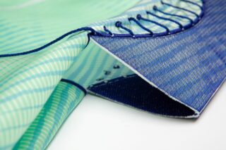 Nahaufnahme eines Stücks hellblau und grün gemusterten Stoffs mit blauen Nähten und Schnürdetails am Rand, das auf einer weißen Oberfläche liegt.