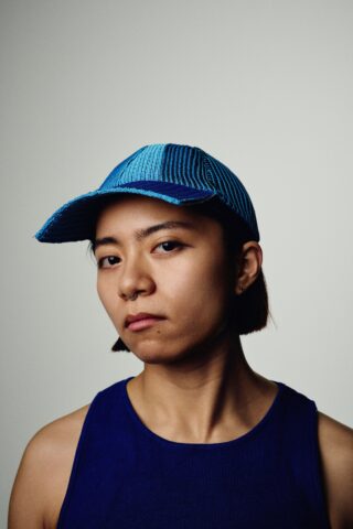 Eine Person mit einer blauen Mütze und einem dazu passenden blauen ärmellosen Oberteil blickt mit neutralem Gesichtsausdruck vor einem schlichten Hintergrund direkt in die Kamera.