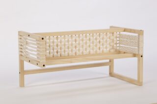 Eine kleine Holzbank mit Seiten- und Rückenlehnen aus gewebtem weißem Kordelzug vor einem schlichten weißen Hintergrund.