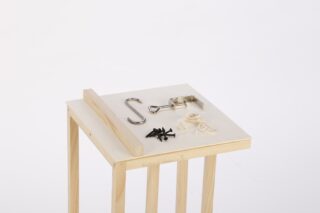 Ein Holzständer zeigt einen Metallhaken, eine Metallklammer, einen Holzstreifen, mehrere Schrauben, ein Bündel Schnur und einige schwarze Plastikteile auf weißem Hintergrund.