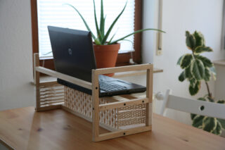 Ein schwarzer Laptop steht auf einem Holzständer auf einem Tisch, im Hintergrund sind eine Topfpflanze und ein Fenster zu sehen.