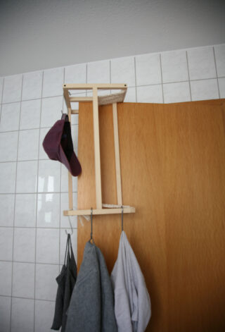 Ein hölzerner Türständer mit einem Besen und einer Mütze, darunter hängen Kleidungsstücke an Haken, befestigt an einer Holztür vor einer gefliesten Wand.