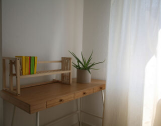 Ein Holzschreibtisch mit einer kleinen Pflanze in einem Topf und einem hölzernen Bücherregal mit mehreren bunten Büchern, neben einem weißen Vorhang, durch den Sonnenlicht hereinströmt.