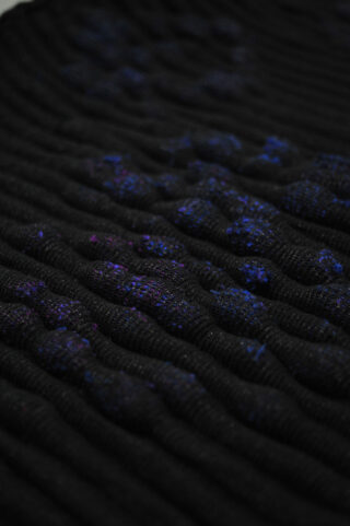 Nahaufnahme eines strukturierten schwarzen Stoffes mit erhabenen, holprigen Mustern und verstreuten blauen und violetten Flecken.