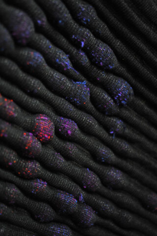 Nahaufnahme eines schwarzen strukturierten Stoffes mit erhabenen Rippen, wobei jeder Abschnitt mit kleinen violetten und roten Punkten gesprenkelt ist.