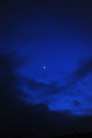 In einem dunkelblauen, teilweise von Wolken bedeckten Nachthimmel sind eine Mondsichel und ein heller Stern zu sehen.