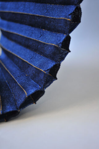 Nahaufnahme eines plissierten blauen Stoffes mit sichtbaren Nähten vor einem schlichten hellen Hintergrund.