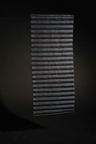 Vertikale, rechteckige Hängeskulptur aus gleichmäßig angeordneten, horizontalen dunklen Lamellen, die vor einem dunklen Hintergrund aufgehängt ist.