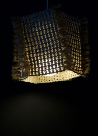 Ein gewebter, quadratischer Lampenschirm mit ausgefransten Rändern wird von innen beleuchtet und wirft einen warmen Schein auf einen dunklen Hintergrund.