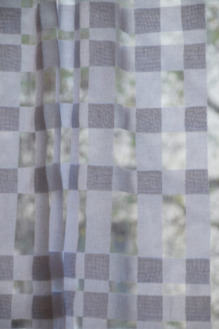 Ein durchsichtiger, karierter Vorhang mit einem weiß-grauen Gittermuster hängt vor einem Fenster, vor dem verschwommenes Grün zu sehen ist.