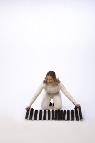 Eine weiß gekleidete Person kniet auf dem Boden und arrangiert ein schwarzes Objekt mit mehreren vertikalen Segmenten auf einem weißen Hintergrund.