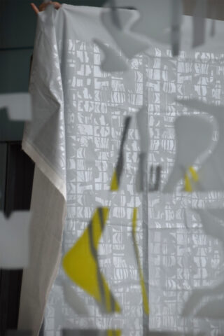 Eine Person hält einen halbtransparenten weißen Stoff mit abstrakten grauen und gelben Mustern hoch, der teilweise durch Milchglas mit geometrischen Designs verdeckt wird.