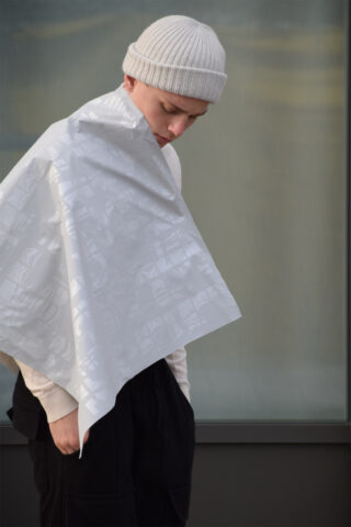 Eine Person mit einer weißen Beaniemütze und einem langärmeligen Hemd steht mit einem gemusterten weißen Tuch über der Schulter da und blickt vor einem unscharfen Hintergrund nach unten.