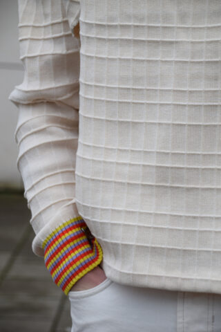 Person, die einen strukturierten cremefarbenen Pullover mit bunt gestreiften Bündchen trägt und mit einer Hand in der Tasche einer weißen Hose steht.