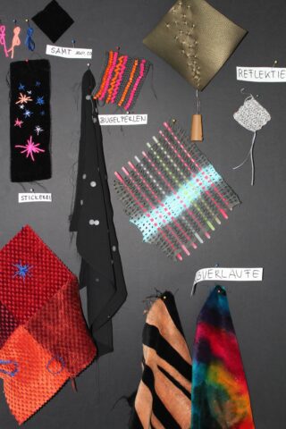 Eine Ausstellung verschiedener Stoffmuster und Stoffproben, jeweils auf Deutsch beschriftet, auf einem schwarzen Hintergrund befestigt.