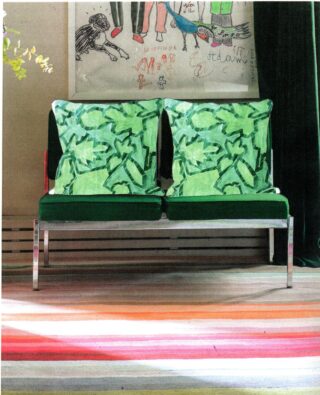 Zwei Stühle mit grünen Kissen stehen nebeneinander an einer Wand, über der auf einem bunt gestreiften Teppich Kinderkunstwerke hängen.