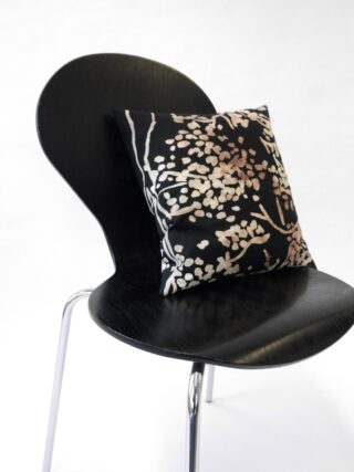 Auf einem schwarzen Stuhl mit Metallbeinen liegt ein quadratisches Kissen mit einem botanischen Muster in Beige und Weiß auf schwarzem Hintergrund.