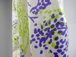 Weißer Stoff mit abstrakten gepunkteten und linierten Mustern in Lila, Grün und Schwarz, der vertikal vor einem schlichten hellgrauen Hintergrund hängt.