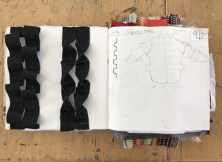 Ein offenes Skizzenbuch liegt auf einem Holztisch und zeigt auf der linken Seite schwarze, gefaltete Papierstreifen und auf der rechten Seite eine technische Umschlagskizze mit Notizen.