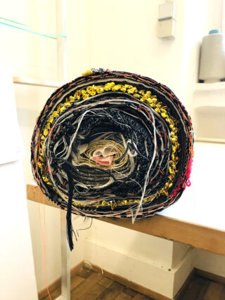 Eine Rolle mit bunten Stoffstreifen und Fäden, die eng miteinander verwoben sind und aufrecht auf einem Holztisch in einem gut beleuchteten Raum stehen.