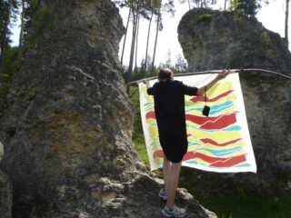 Eine Person in einem schwarzen Outfit hält einen bunten Stoff mit abstraktem Muster, der über einen Stock zwischen zwei großen Felsformationen im Freien drapiert ist.