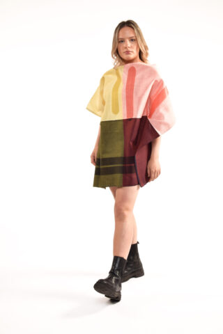 Vor einem schlichten Hintergrund steht eine Person, die ein kurzes, locker sitzendes Kleid mit gelben, rosa und grünen Farbblöcken und schwarze Stiefel trägt.