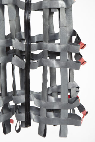 Eine Nahaufnahme eines grauen Gurtzeugs mit ineinander verschlungenen Riemen, einem schwarzen Reißverschluss in der Mitte und roten Aufreißlaschen an der Seite vor einem weißen Hintergrund.