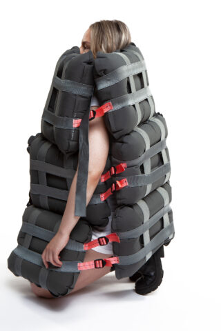 Eine Person trägt einen großen, gepolsterten, grauen Anzug aus rechteckigen Kissen, die mit grauen und roten Verschlüssen zusammengebunden sind und den größten Teil ihres Körpers bedecken.