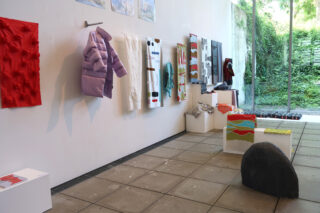 Kunstinstallation mit bunten Textilien, einer lila Pufferjacke und abstrakten Objekten, die entlang einer weißen Galeriewand mit großen Fenstern mit Blick ins Grüne ausgestellt sind.