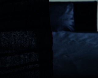 Ein durchsichtiger, dunkler Vorhang verdeckt teilweise ein ordentlich gemachtes Bett mit dunkelblauer Bettwäsche und Kissen in einem schwach beleuchteten Raum.