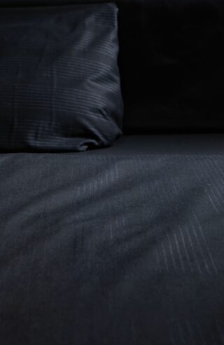 Eine Nahaufnahme eines dunkelfarbigen Bettlakens und Kissens mit dezentem Streifenmuster, die einen Teil der Bettoberfläche und die untere Hälfte des Kissens zeigt.