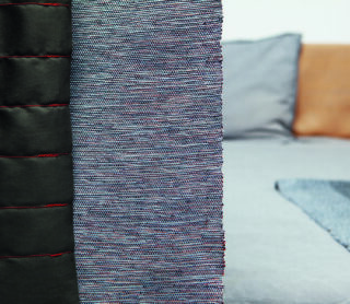 Nahaufnahme eines strukturierten Gewebes neben einem schwarzen Material mit roten Nähten; im Hintergrund sind verschwommene Kissen und ein Bett zu sehen.