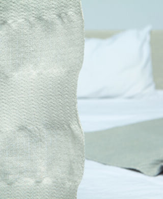 Eine Nahaufnahme eines strukturierten hellen Vorhangs mit einem ungemachten Bett, einem weißen Kissen und einer gefalteten Decke im Hintergrund.