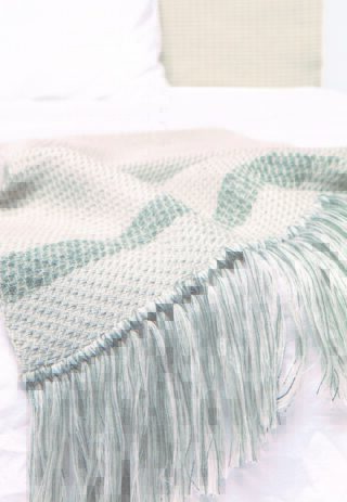 Eine gewebte Decke mit grünen und weißen Streifen und Fransenrändern liegt über einem weißen Bett.