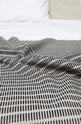 Eine Nahaufnahme einer schwarz-weiß karierten Decke mit sichtbaren Falten, ausgebreitet auf einem Bett mit weißen Laken und einem Kissen im Hintergrund.