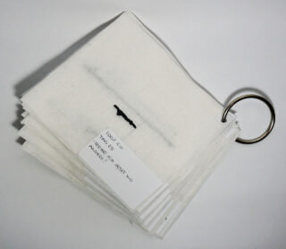 Ein Stapel weißer Stoffmuster, die durch einen Metallring zusammengehalten werden. Am oberen Muster ist ein Etikett mit handschriftlichen Notizen angebracht.