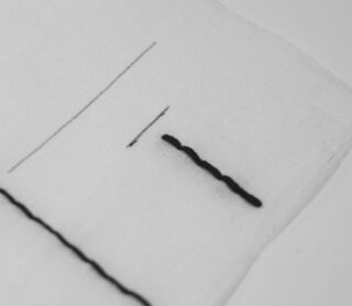Eine Nahaufnahme von weißem Stoff mit schwarzen Fadenstichen und geraden schwarzen Linien, die einen Vergleich zwischen handgenähten und gezeichneten Linien zeigt.