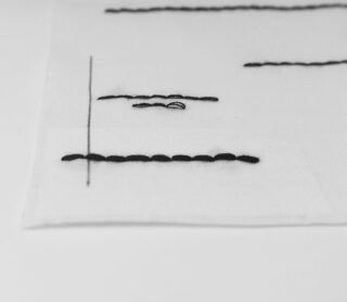 Ein weißes Stück Stoff mit mehreren geraden und wellenförmigen schwarzen Nähten, die horizontal und vertikal verlaufen.