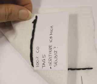 Eine Hand hält ein Stück weißen Stoff mit schwarzen Nähten und einem Papieretikett mit der handgeschriebenen Aufschrift „100 % CO TAG 5 – IRRITIERE ICH MICH SELBST?“.