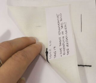 Eine Hand hebt die Ecke des weißen Stoffes mit der schwarzen Naht an und gibt den Blick auf ein Etikett frei, auf dem handgeschriebener deutscher Text und die Aufschrift „100 % CO“ stehen.