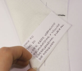 Eine Hand hebt ein Stück weißen Stoff an und gibt den Blick auf eine Karte frei, auf der „100 % CO“ steht und ein handgeschriebener deutscher Text steht.